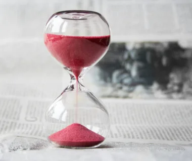 Close-up of a transparent hourglass with pink sand flowing, placed on a newspaper background.